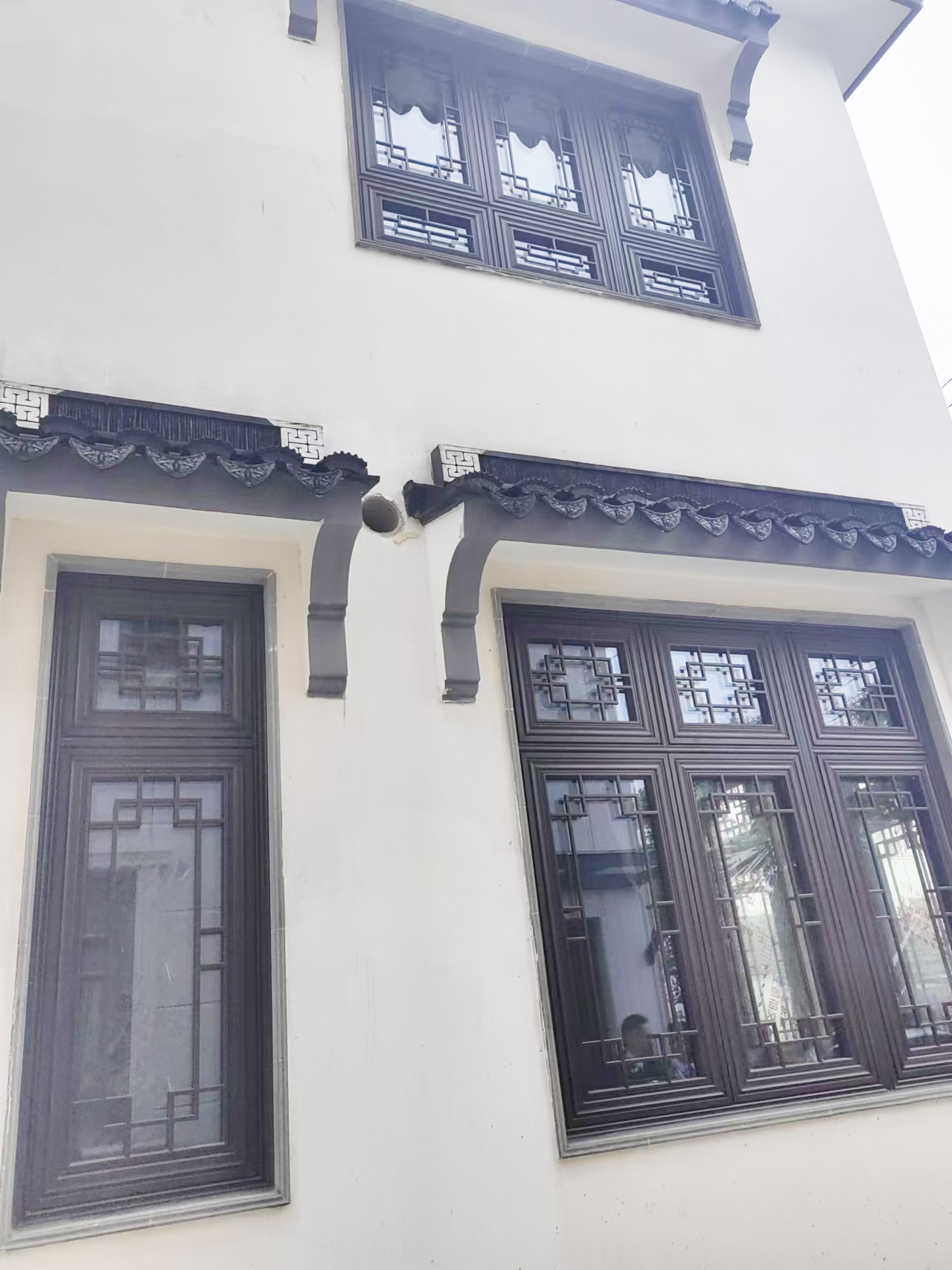 这张图片展示了白墙建筑外立面的局部特写，核心主体为深灰色新中式铝合金门窗。窗户采用经典的中式回纹格栅设计，上方搭配带有瓦片纹理和回纹装饰的仿古屋檐，尽显江南水乡